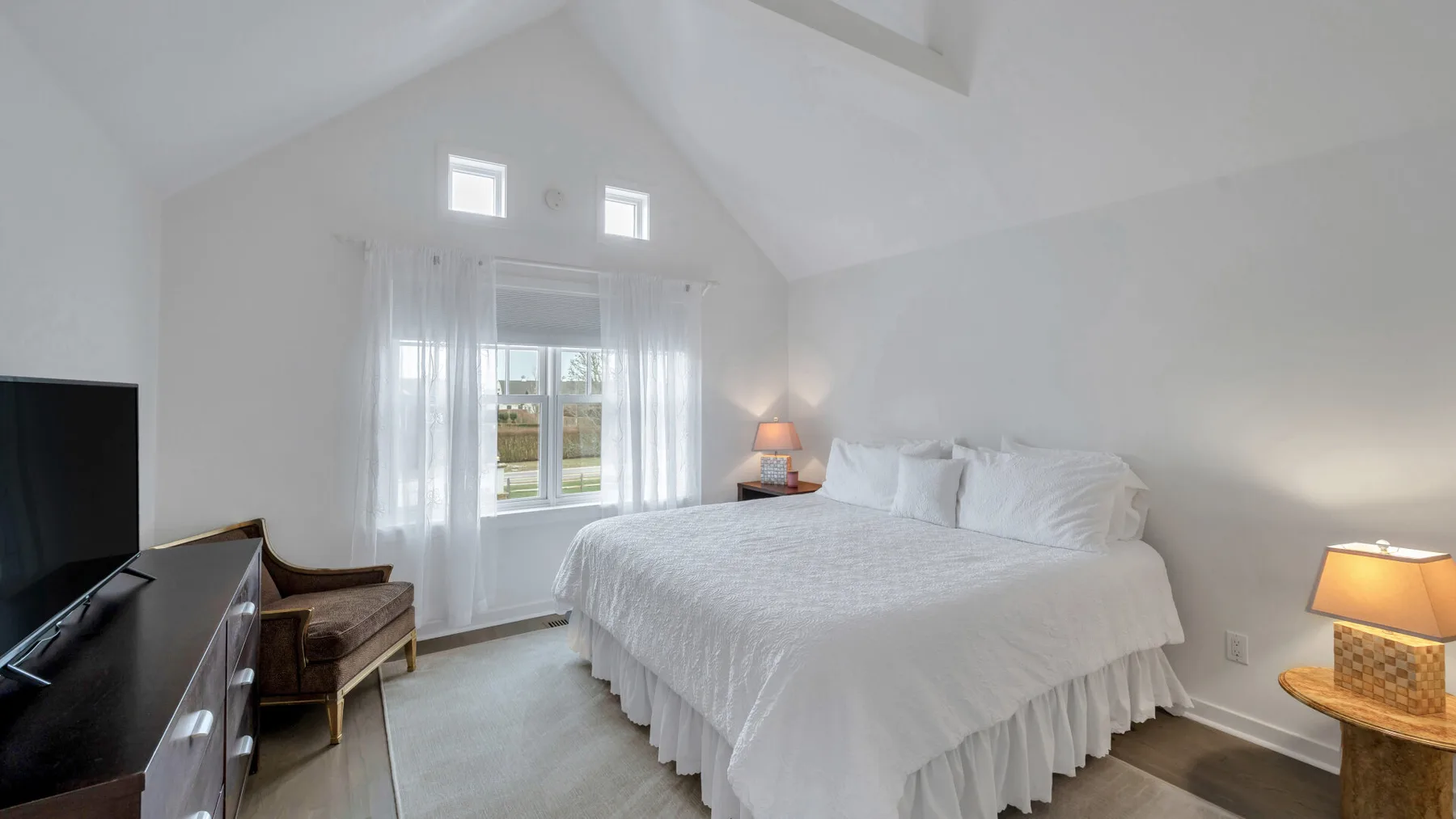 Un dormitorio luminoso con una cama grande, ventanas altas y un televisor.