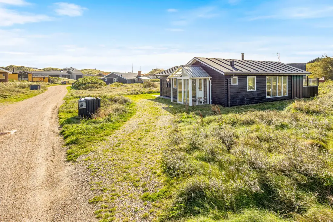 Una casa oscura está junto a un camino de arena entre dunas cubiertas de hierba.