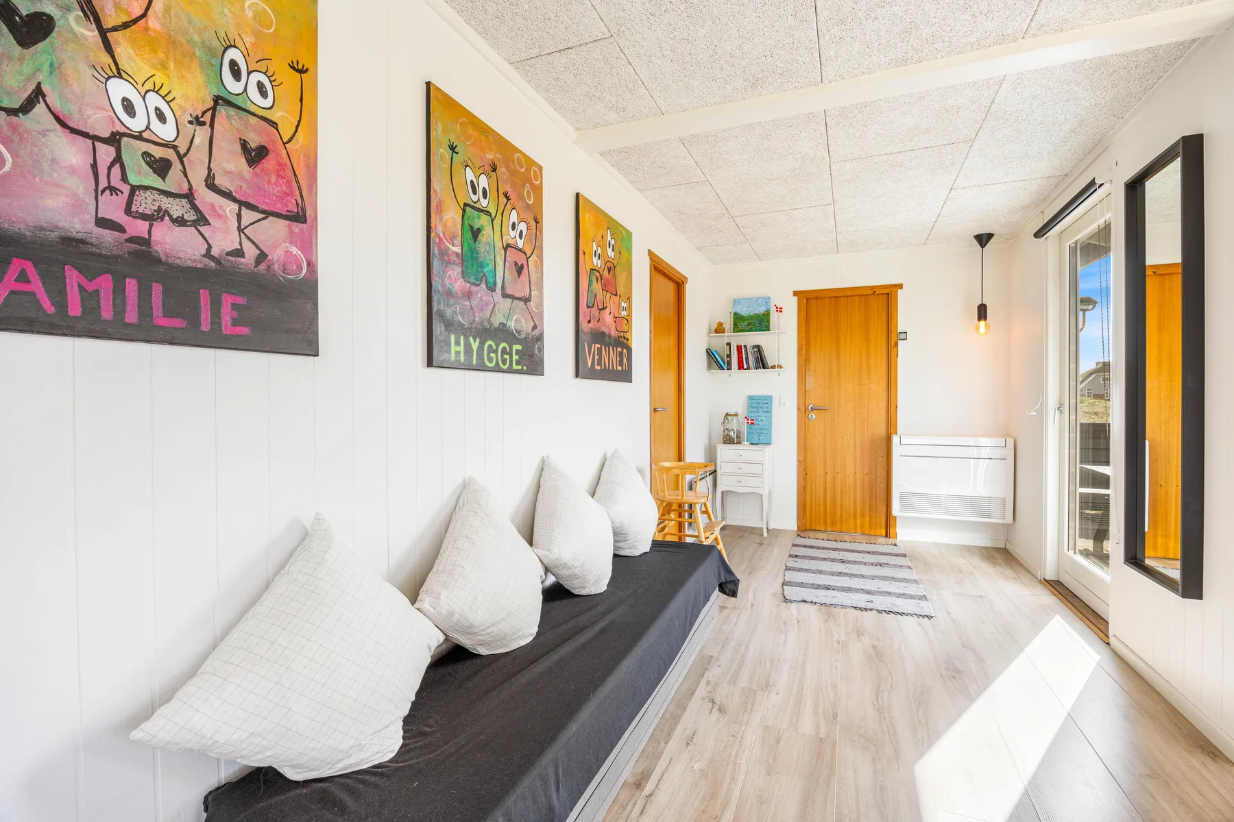 Una sala de estar luminosa tiene un diván oscuro bajo cuadros coloridos.