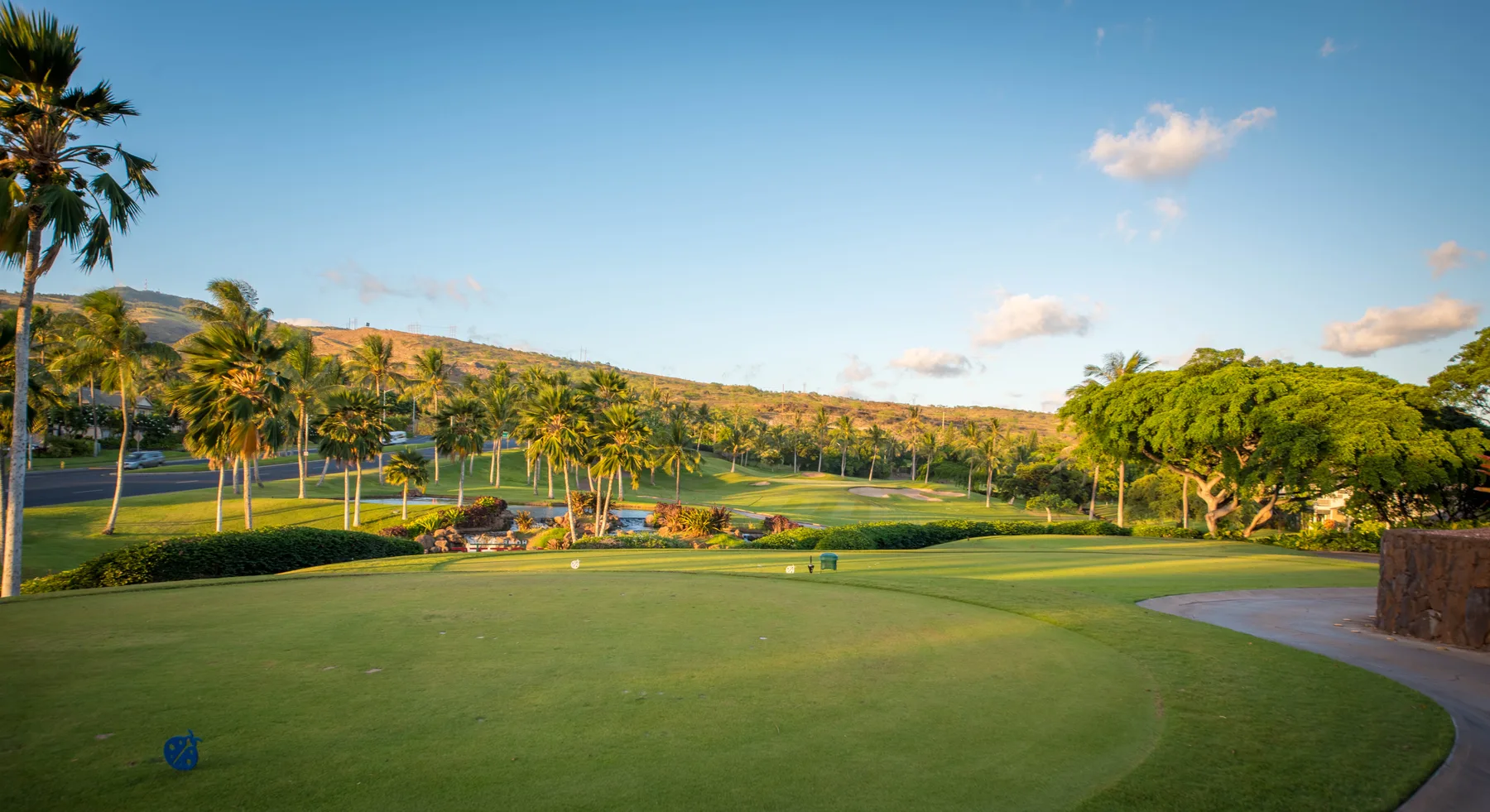 Un campo de golf ajardinado se extiende en primer plano, con palmeras y colinas al fondo.