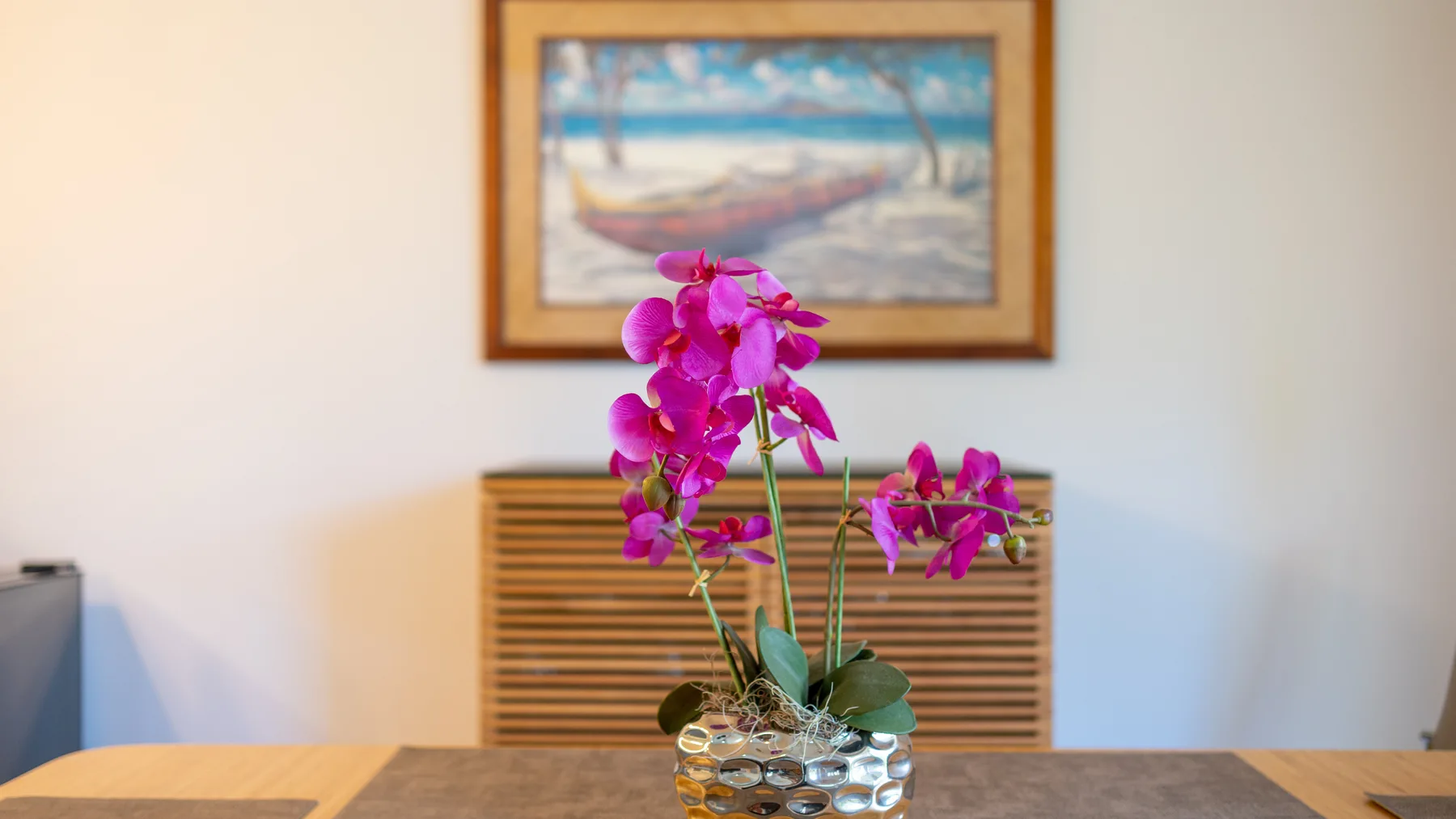 Una orquídea en una maceta está sobre una mesa frente a un cuadro enmarcado.