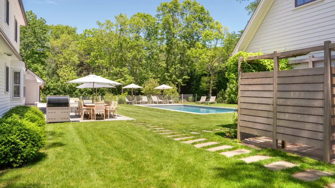Un patio con césped, piscina, asientos al aire libre y árboles.