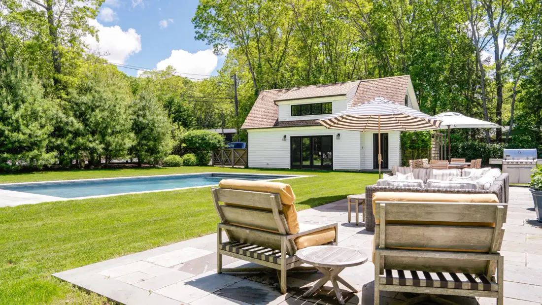 A patio with outdoor seating overlooks a swimming pool beside a white house.