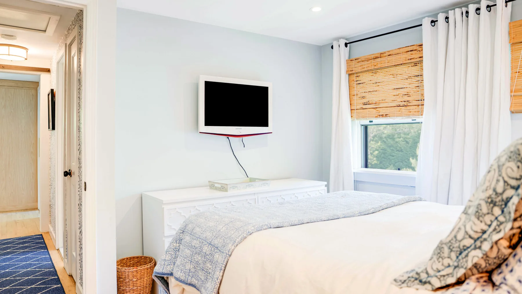 Un dormitorio luminoso con una cama tendida, un televisor en la pared y una ventana con cortinas.