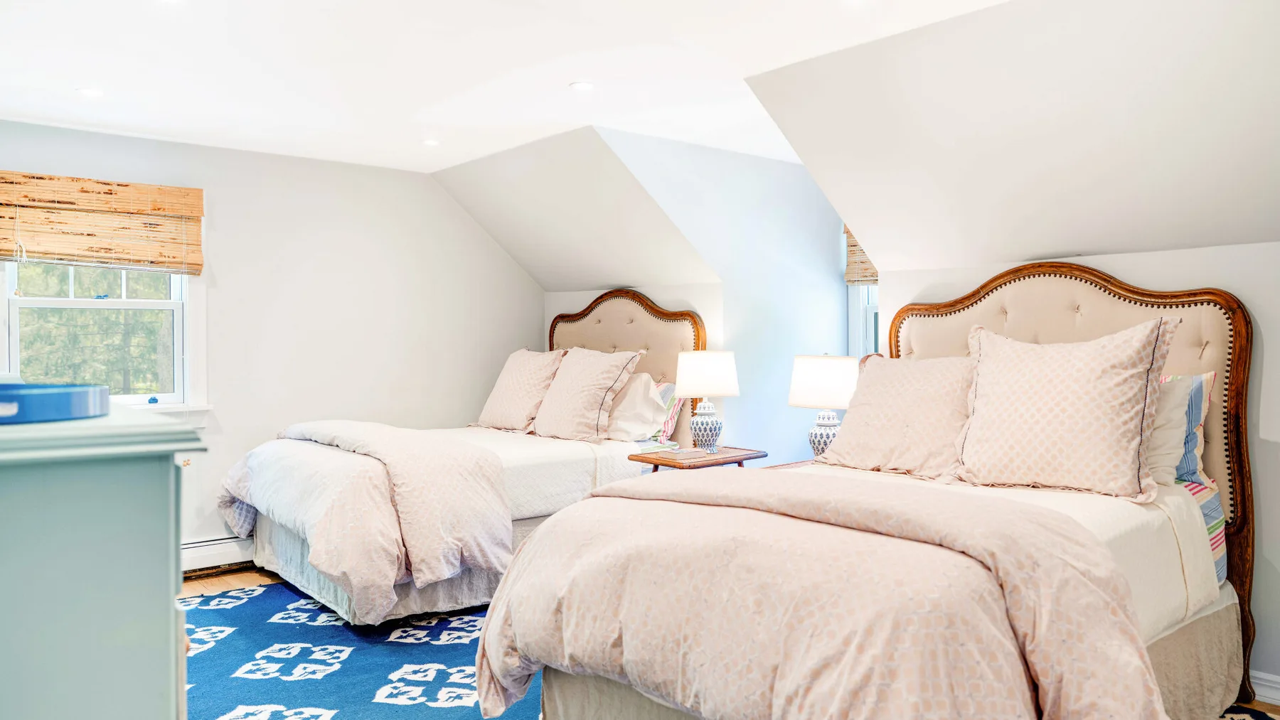 Un dormitorio luminoso con dos camas, ropa de cama clara y una alfombra azul estampada.