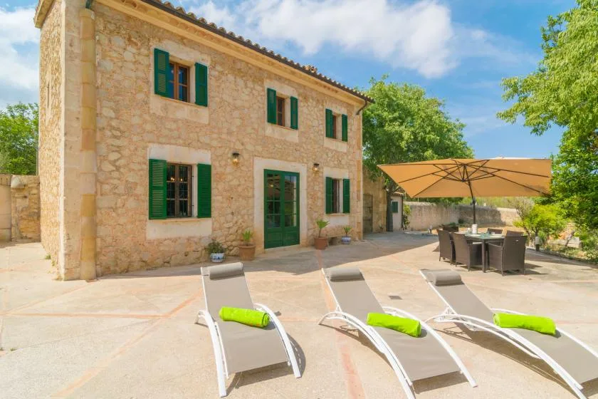 A stone house opens onto a sunny patio with loungers and a shaded dining area.