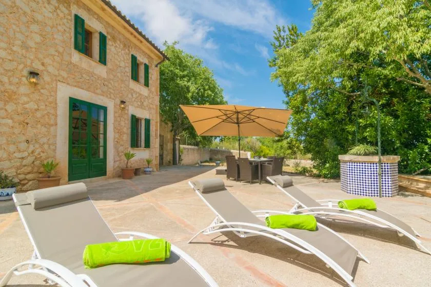 A sunny patio with reclining loungers, a large umbrella, and leafy trees.