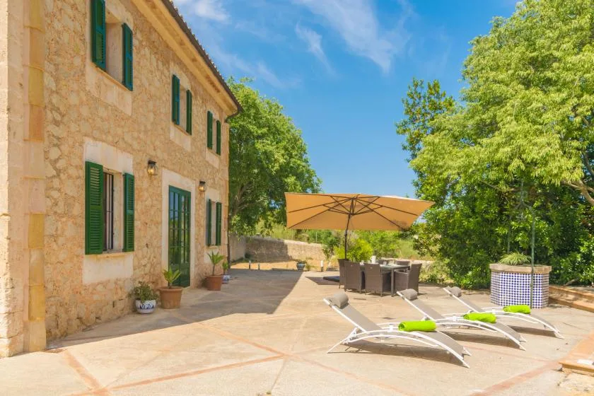 A stone house opens onto a sunny patio with loungers and a large umbrella.
