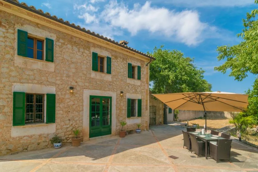 A stone house opens onto a sunlit patio with outdoor seating and a large umbrella.
