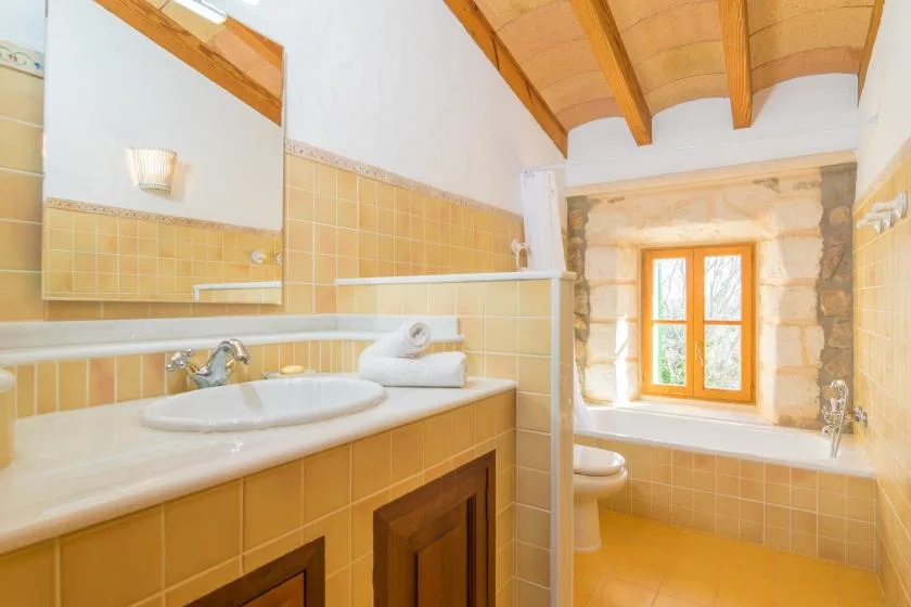 A tiled bathroom with a sink, bathtub, and window beneath exposed wooden beams.