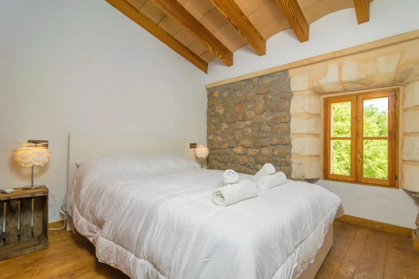 A bedroom with a white bed, exposed wooden ceiling beams, and a stone wall.