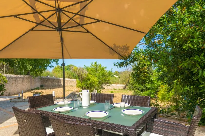 An outdoor dining table sits beneath a large patio umbrella amid greenery.