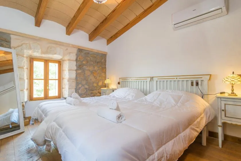 A bedroom with white bedding, wooden ceiling beams, and a stone accent wall.