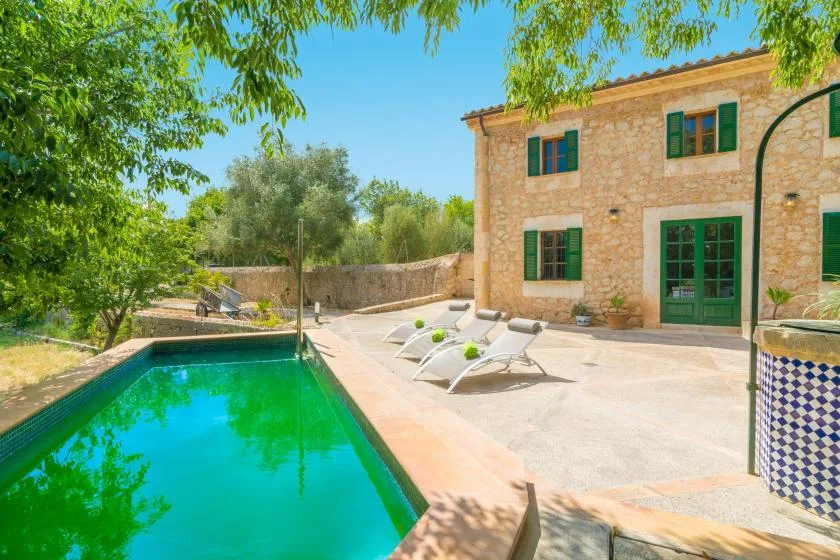 A swimming pool beside a stone house, with lounge seating on the paved patio.