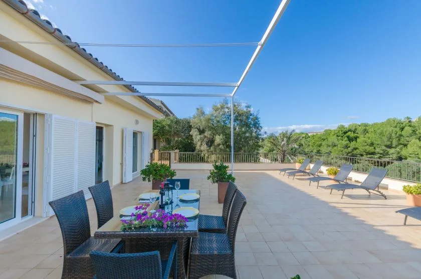 Una terraza soleada con comedor, tumbonas y vistas a una arboleda.