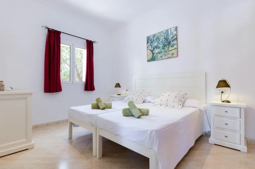Un dormitorio luminoso con ropa de cama blanca, cortinas rojas y un cuadro colorido sobre las camas.