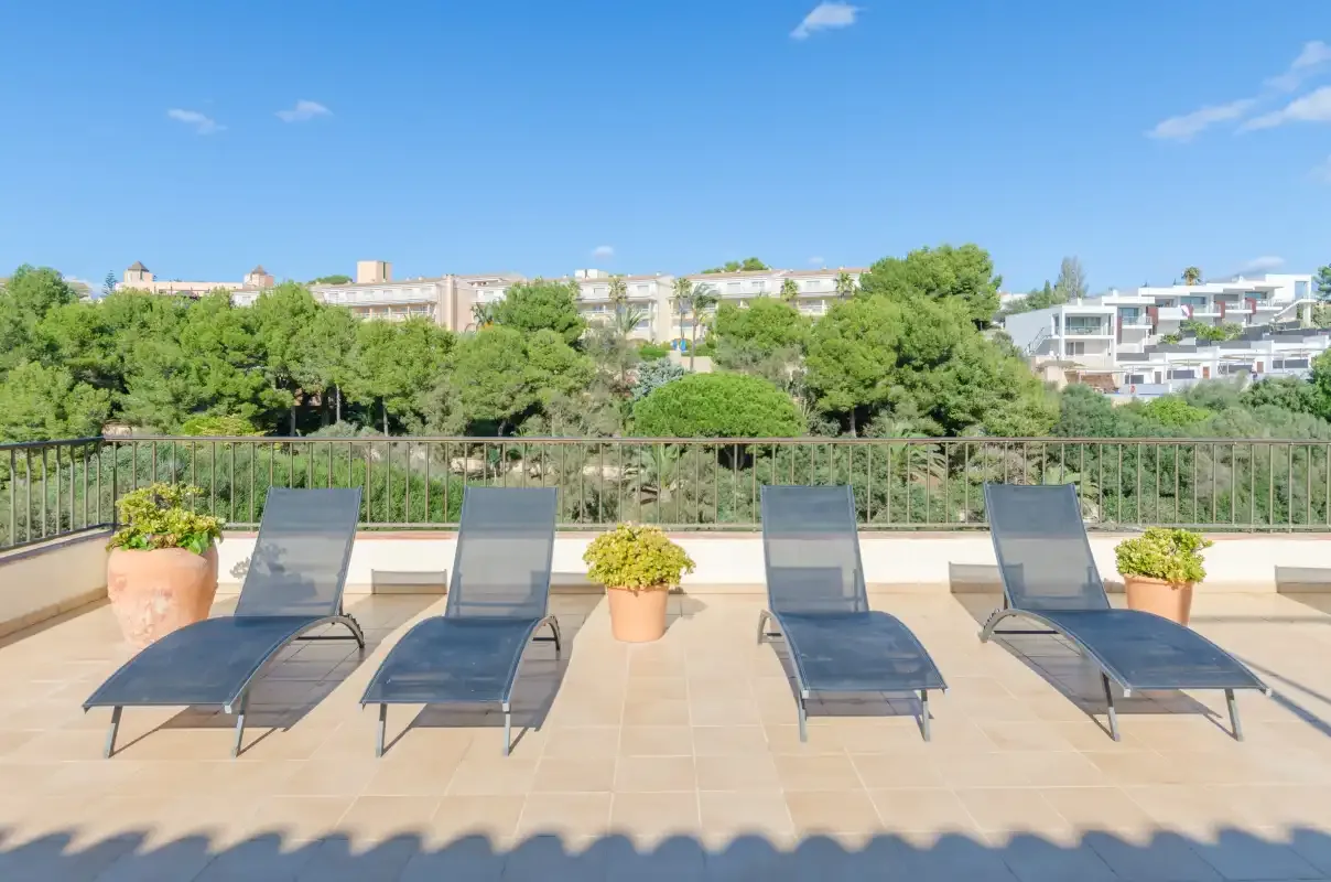 Una terraza soleada con asientos reclinables y plantas en macetas da a árboles y edificios.