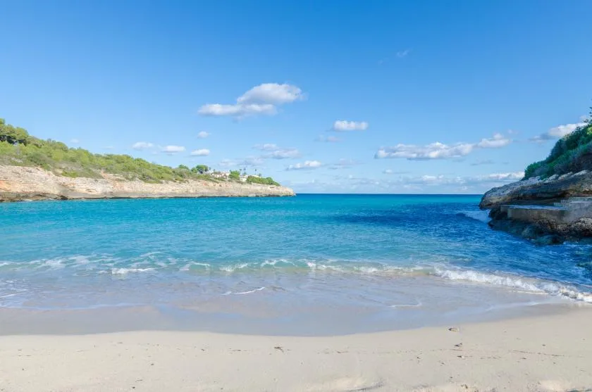 Una playa de arena bordea una bahía azul tranquila entre costas rocosas cubiertas de vegetación.