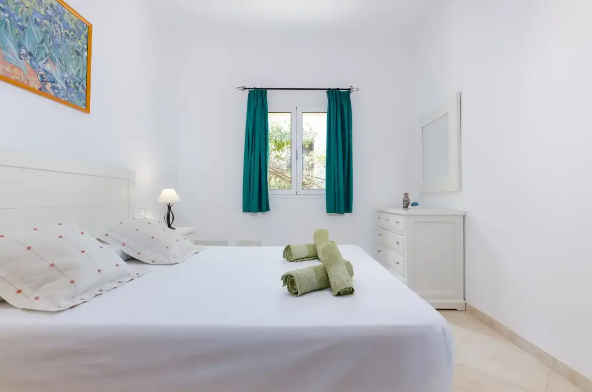 Dormitorio luminoso con una cama, cortinas turquesas y una cómoda blanca.