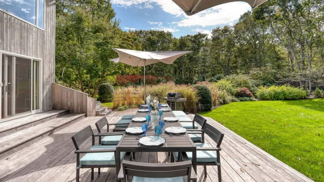 An outdoor dining area on a wooden deck overlooks a green lawn and trees.