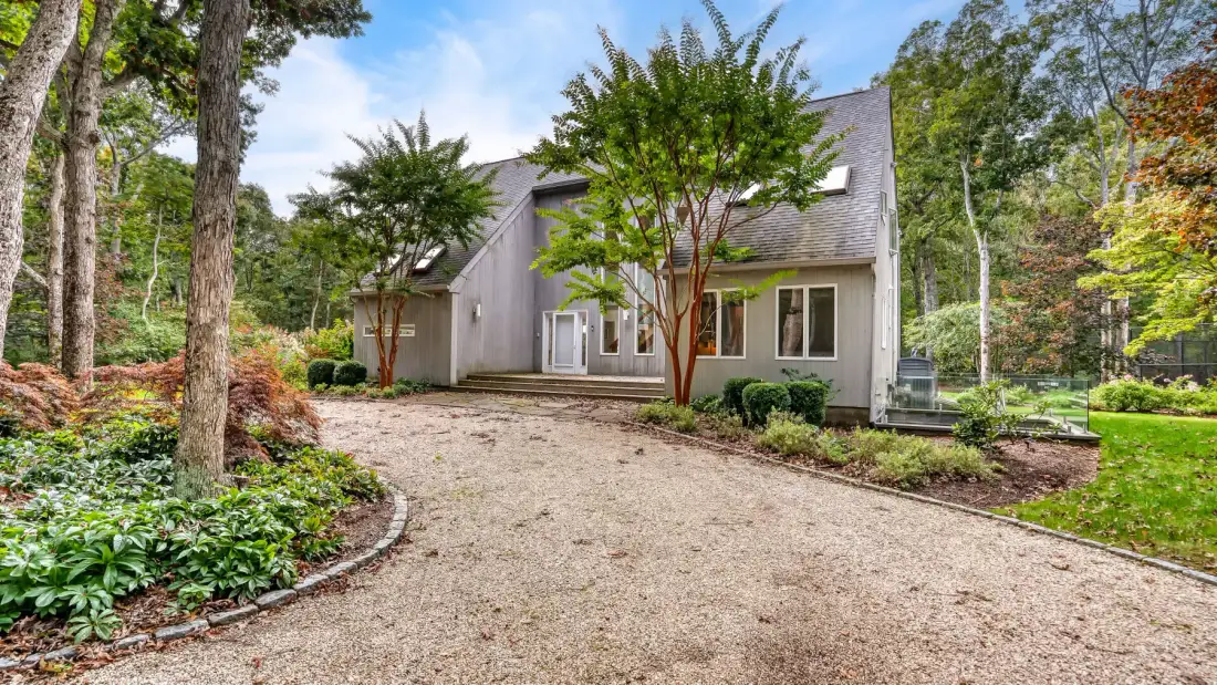 A house stands beyond a curved gravel driveway, surrounded by trees and garden plants.