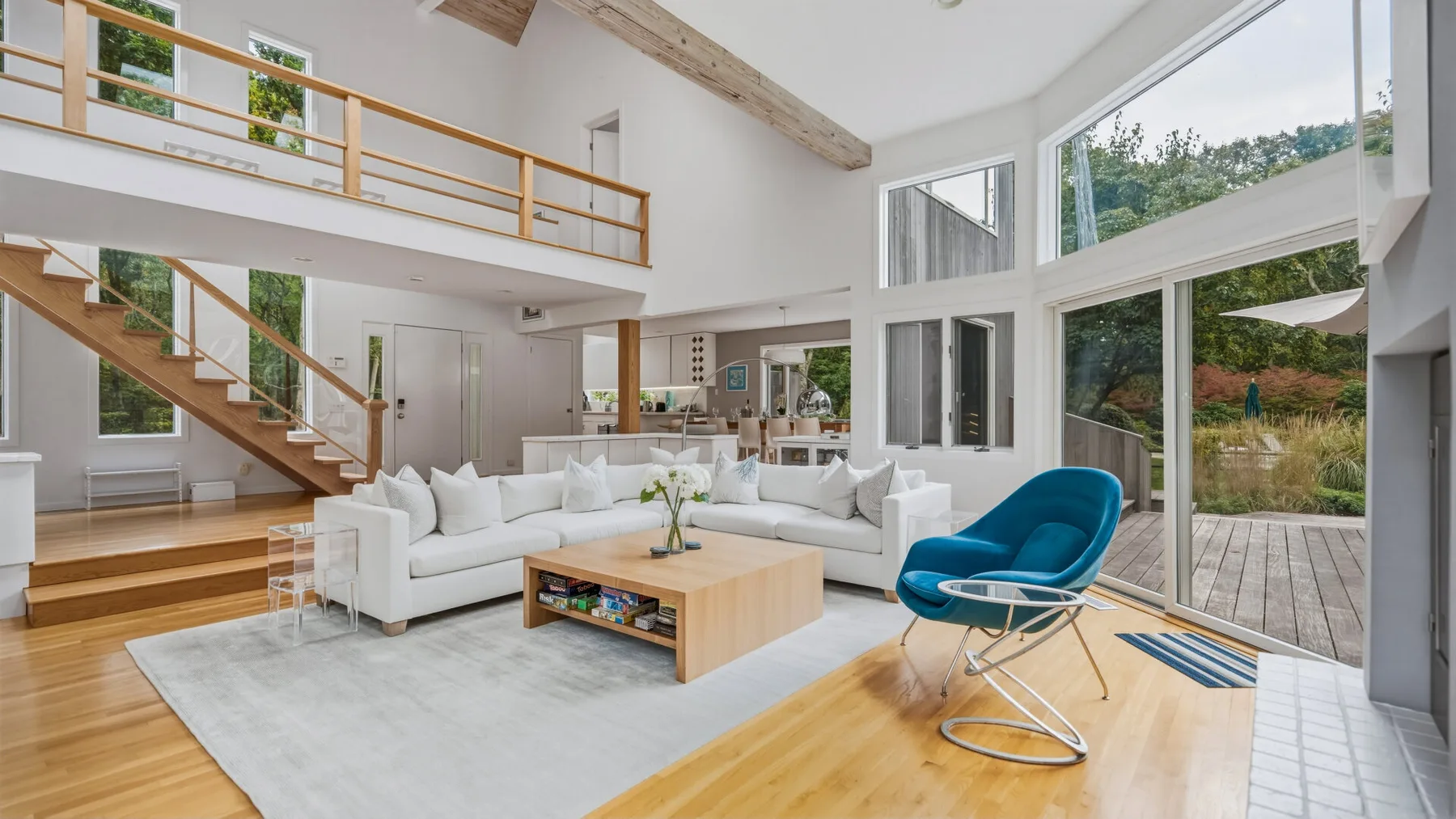A bright living room with white seating, large windows, and a wooden staircase.