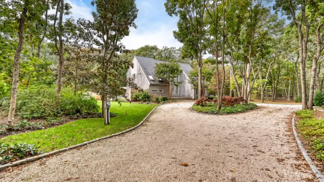 A house stands beyond a curving gravel driveway amid trees and landscaped greenery.