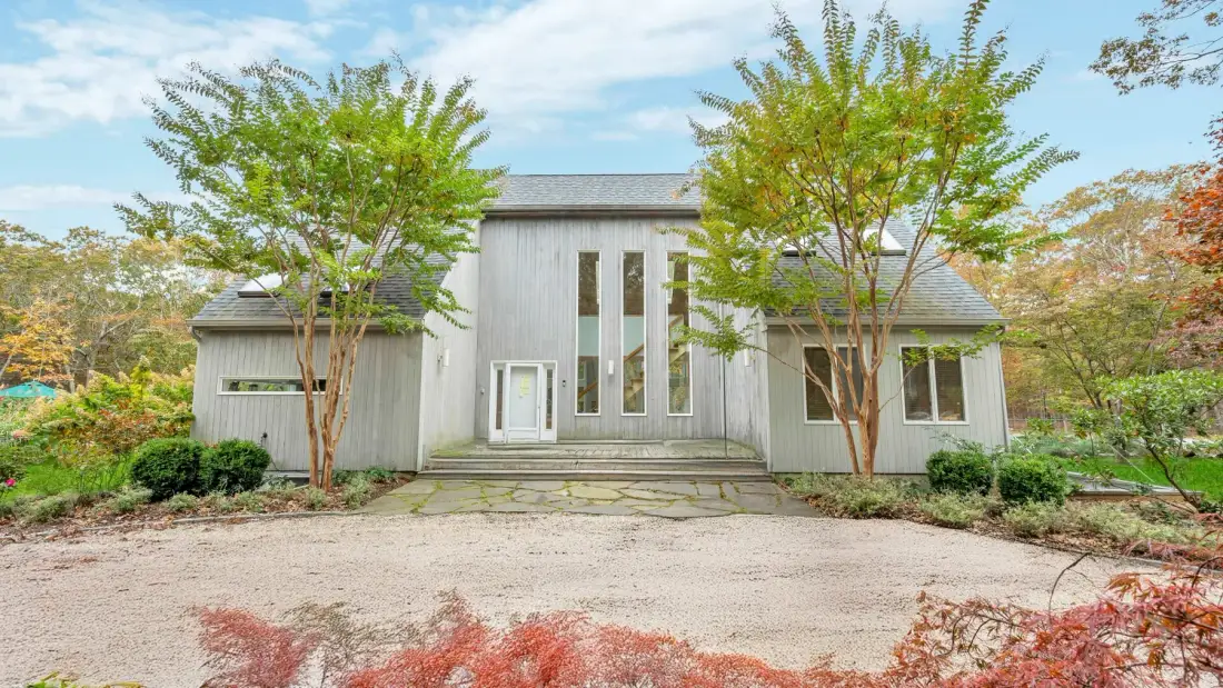A modern house stands beyond a gravel driveway, framed by leafy trees.