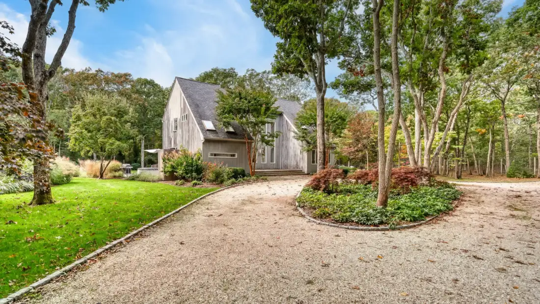 A small house sits beside a gravel driveway in a wooded garden.