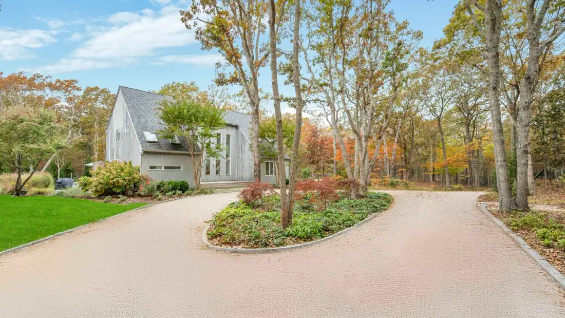 A paved driveway curves around a landscaped island beside a house and trees.
