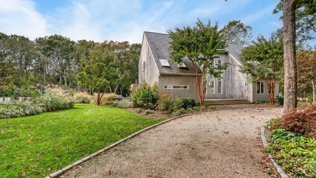 A gravel driveway curves through landscaped grounds toward a gray house.