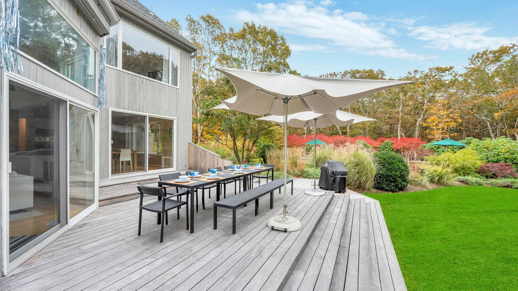 A wooden deck with outdoor dining furniture and a large shade umbrella overlooks a green lawn.