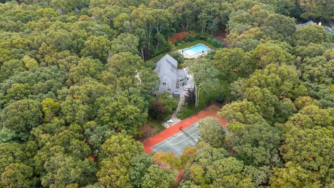 An aerial view shows a house, swimming pool, and tennis court amid dense trees.