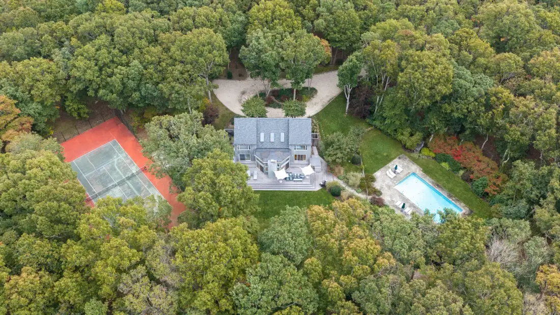 An aerial view shows a house, tennis court, and swimming pool among trees.