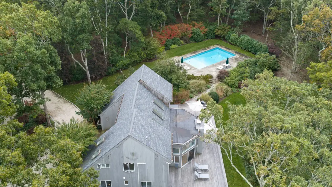 An aerial view shows a gray house surrounded by trees, with a swimming pool nearby.