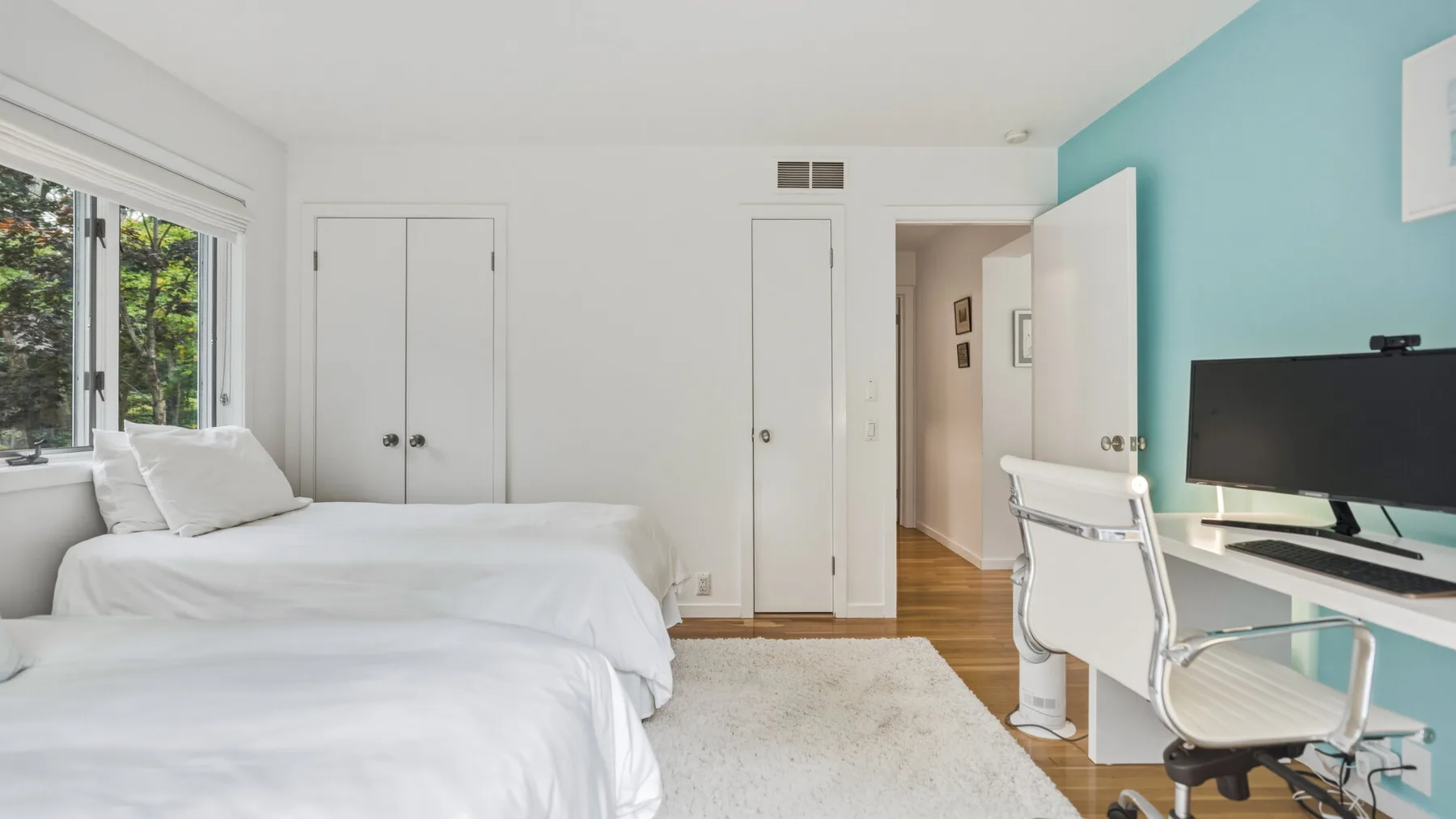 A bright bedroom with white beds, a large window, and a desk beside a turquoise wall.