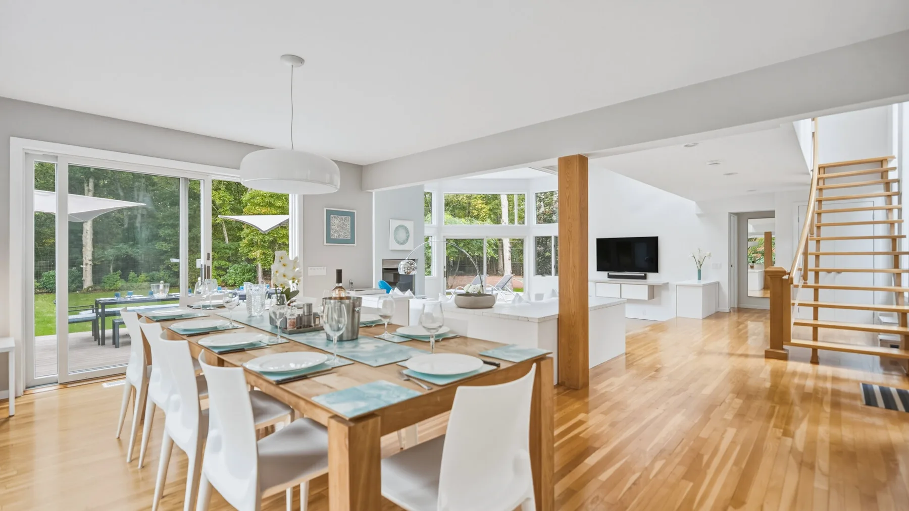 A bright open-plan dining and living area has large windows and a wooden dining table.