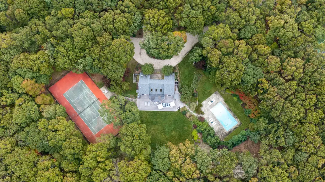 An aerial view of a wooded property with a tennis court and swimming pool.