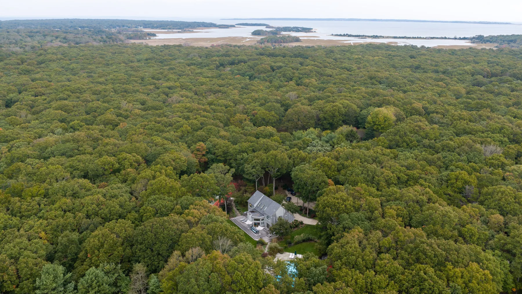 An aerial view shows a house surrounded by dense woodland, with water in the distance.