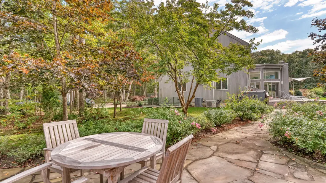 A stone patio with outdoor seating overlooks a landscaped garden and a house.