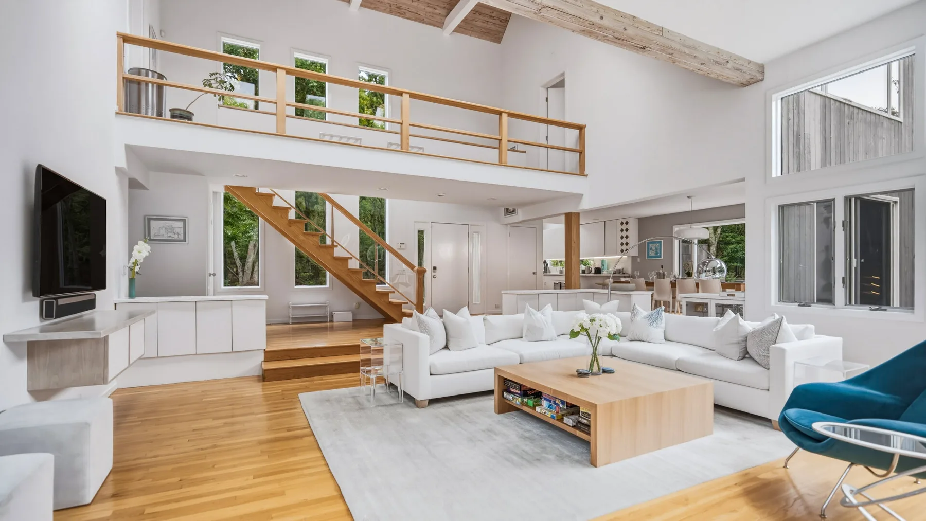 A bright open-plan living room has a staircase, upper gallery, and white seating.