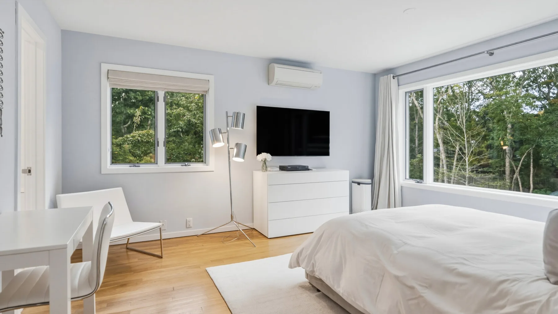 A bright bedroom with a large bed, television, and windows overlooking trees.