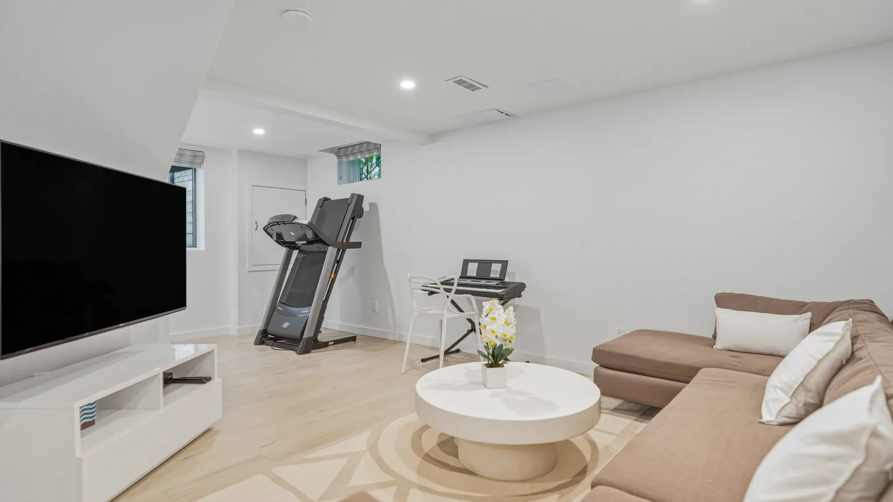 A bright living room with a television, treadmill, and seating around a round coffee table.