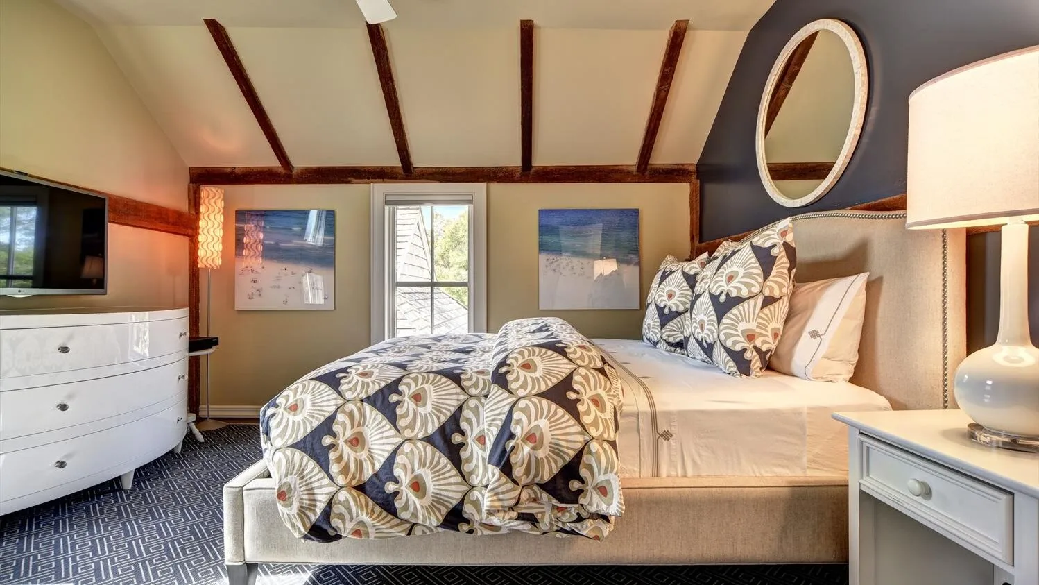 A bedroom with a patterned bed, a window, and dark ceiling beams.