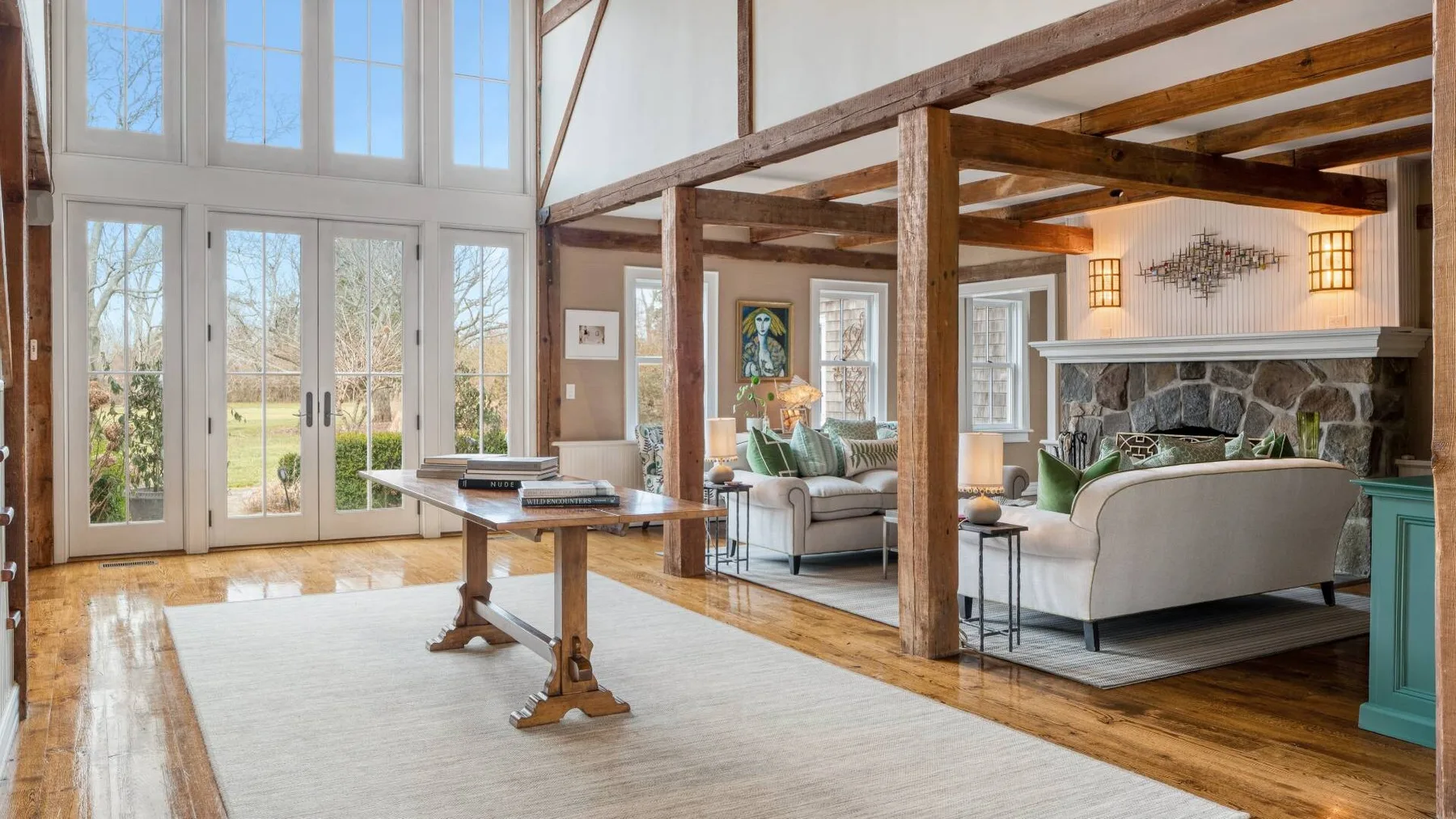A bright living room with tall windows, timber beams, and seating around a stone fireplace.