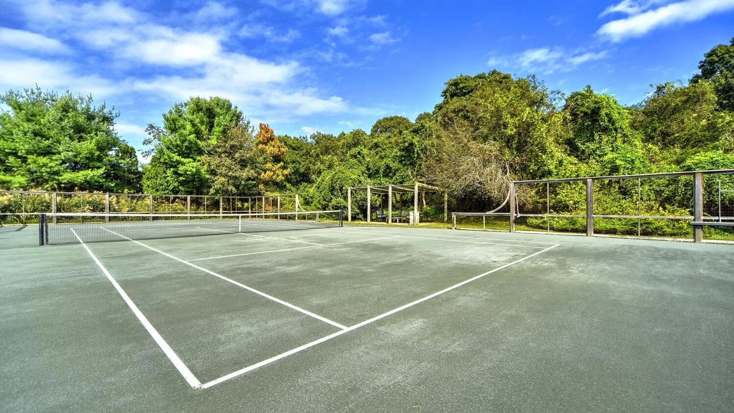 An outdoor tennis court with painted boundary lines and trees beyond the fence.