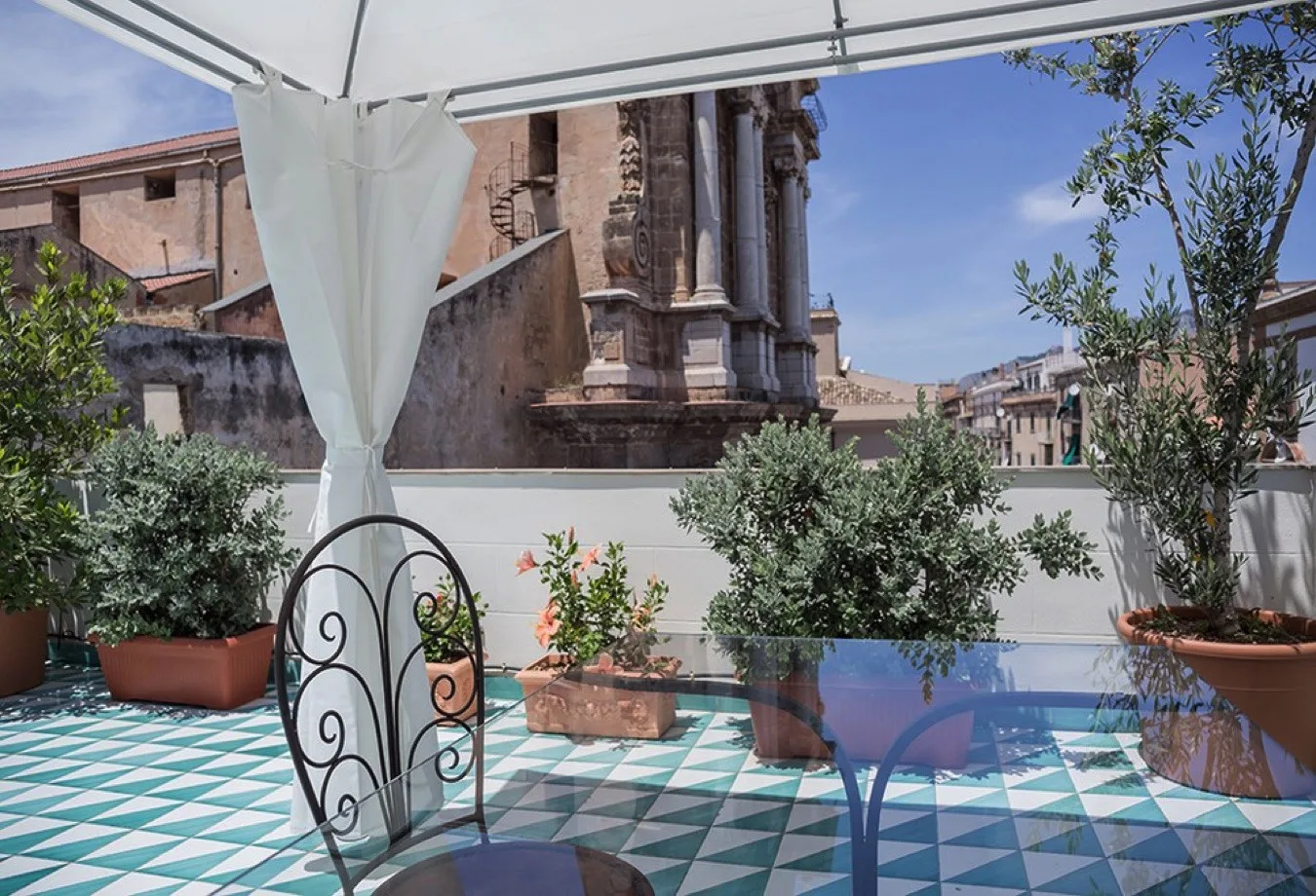 Una terraza exterior con suelo de baldosas, plantas en macetas y edificios históricos a la vista.