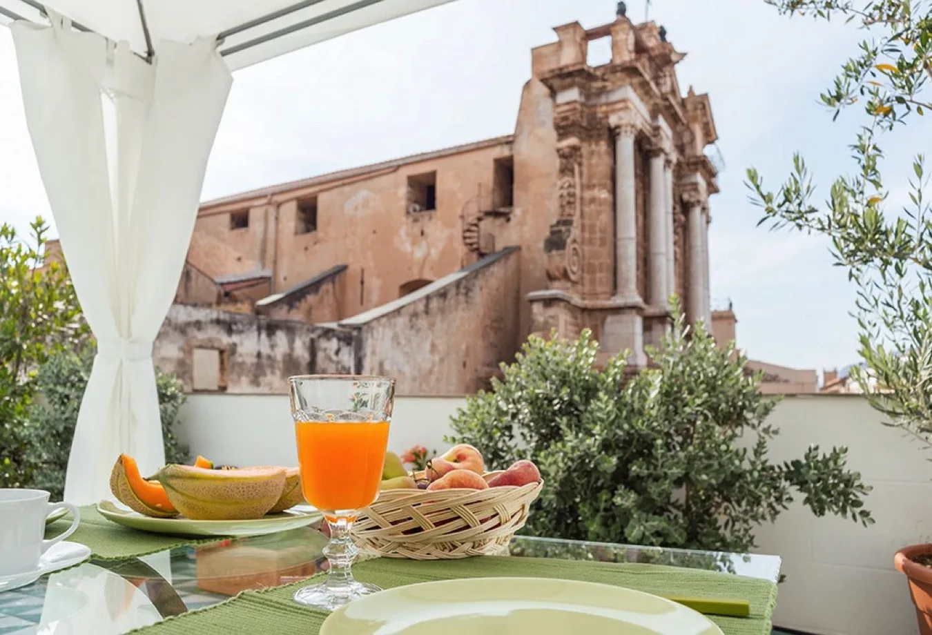 Una mesa de comedor al aire libre con fruta y zumo da a un edificio antiguo de piedra.