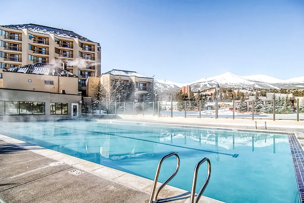 An outdoor swimming pool sits beside a resort building with snowy mountains beyond.