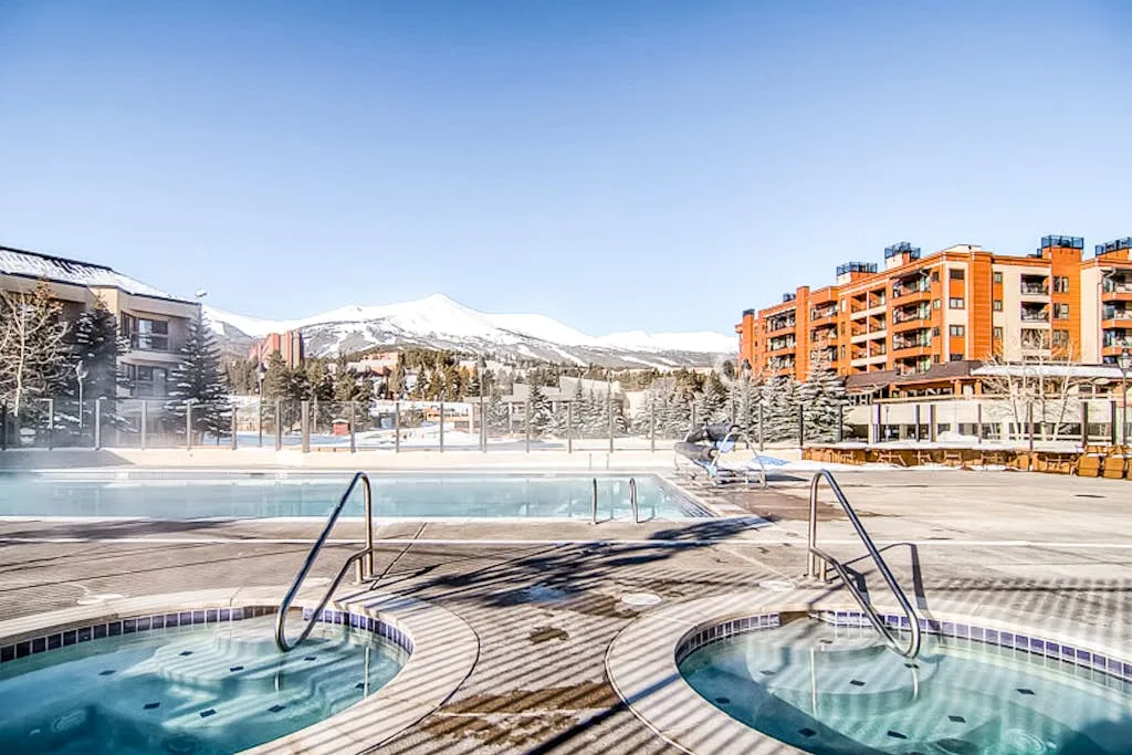 Outdoor pool and round hot tubs overlook snowy mountains and nearby buildings.